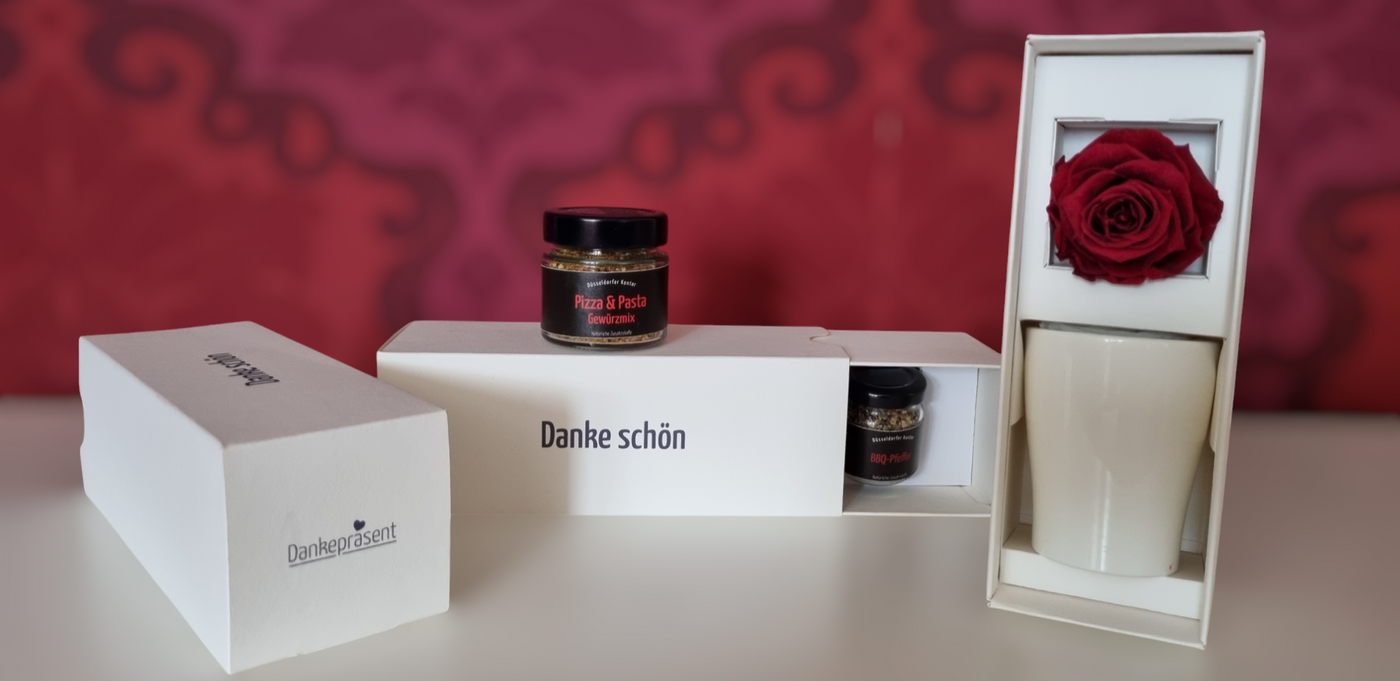 Hochwertige Dankepräsent-Boxen mit „Dankeschön“-Veredelung vor einer edlen roten Ornament-Tapete – stilvolle Mitarbeiter- und Kundengeschenke vom Düsseldorfer Kontor für das gesamte Rheinland und bundesweit.
