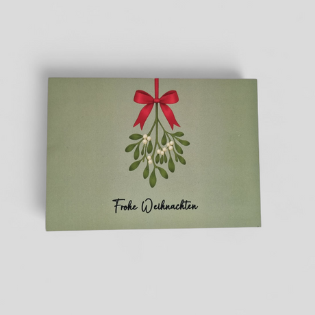 Eine feierliche Weihnachtskarte auf salbeigrünem Hintergrund mit einem Mistelzweig, einer roten Schleife und dem Text „Frohe Weihnachten“.  Das Bild steht für die bundesweite Bereitstellung von hochwertiger Mitarbeiterbindung und authentischem Employer Branding durch Dankepräsent, um Unternehmen dabei zu unterstützen, zum Jahresausklang „Danke“ zu sagen.
